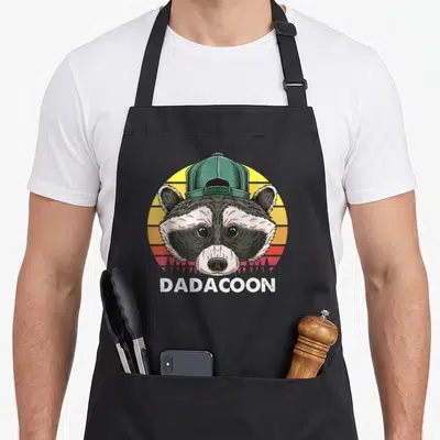 Trash father day apron - Cartoon raccoon face in green cap with DADACOON text on retro sunset — shown on a black cotton canvas apron against a white background