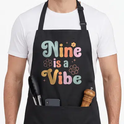 9th boho apron - Stacked Nine is a Vibe text with colorful flowers and stars — shown on a black cotton canvas apron against a white background