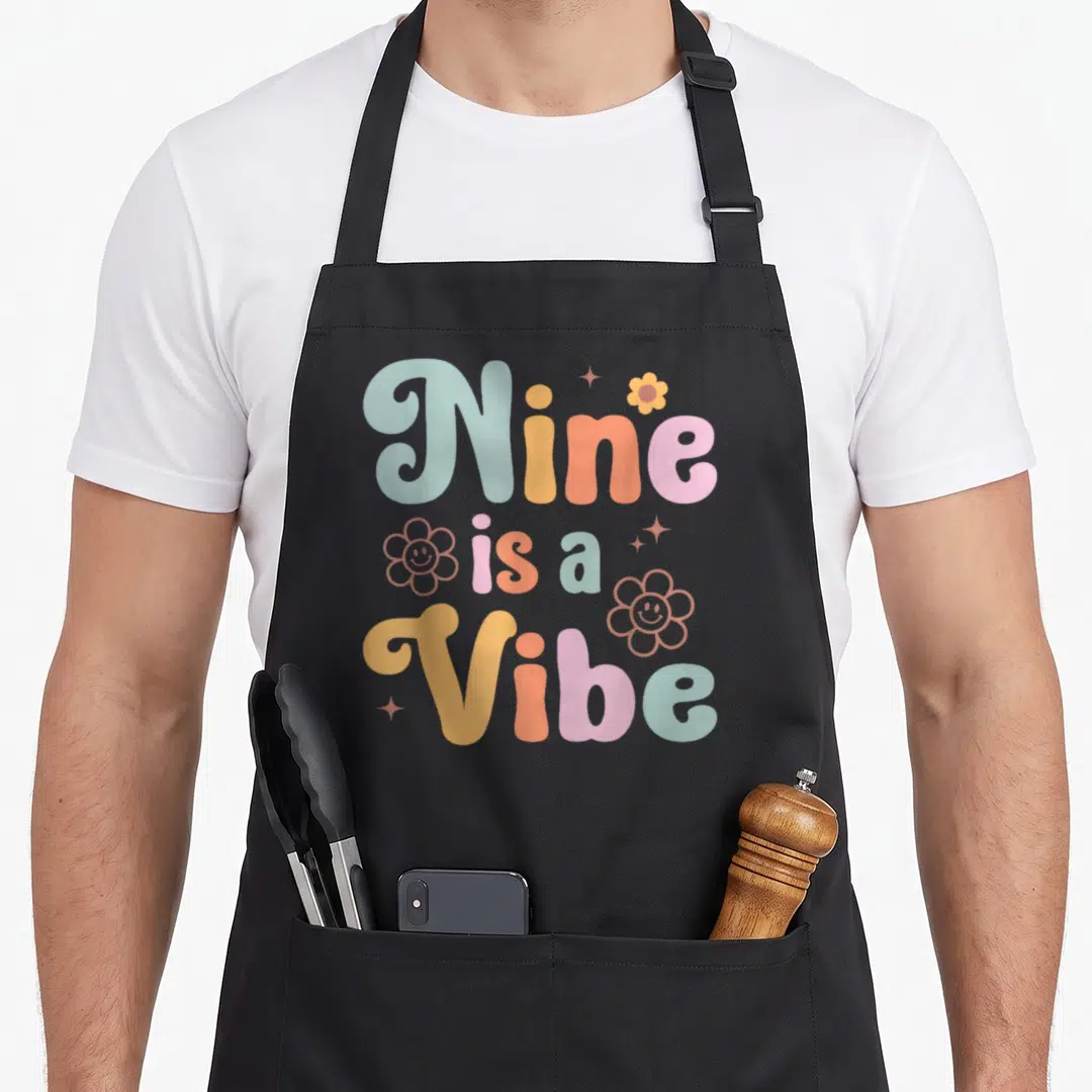 9th boho apron - Stacked Nine is a Vibe text with colorful flowers and stars — shown on a black cotton canvas apron against a white background