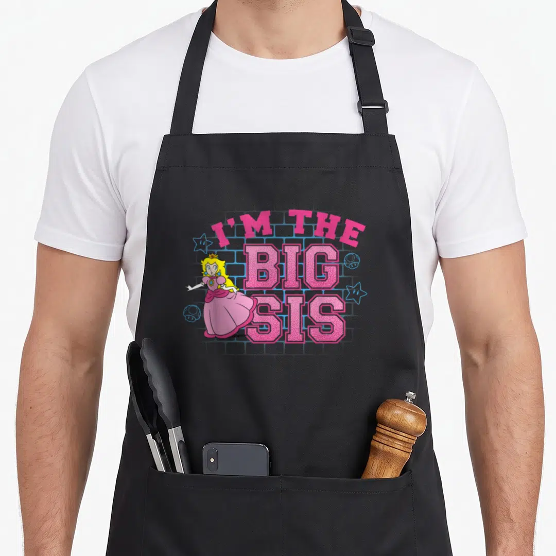 Sis peach apron - Princess Peach I'M THE BIG SIS text pink blue yellow cartoon brick wall — shown on a black cotton canvas apron against a white background