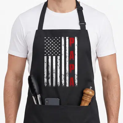 Proud father day apron - Distressed USA flag with vertical red PAPA text on white stripes — shown on a black cotton canvas apron against a white background