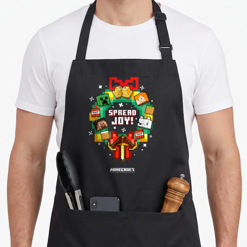 Crown christmas apron - Pixelated Minecraft wreath with spread joy text and character icons — shown on a black cotton canvas apron against a white background