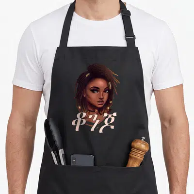 Amharic beautiful apron - Cartoon woman portrait with gold hair beads and Amharic text ቆን in off-white across chest — shown on a black cotton canvas apron against a white background