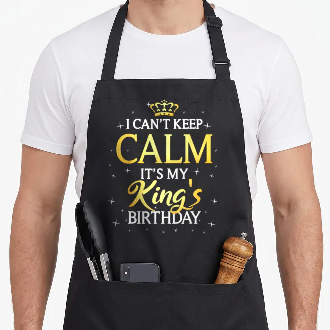 Birthday bday kings apron - Gold crown above white and gold text I Can't Keep Calm It's My King's Birthday with sparkles — shown on a black cotton canvas apron against a white background