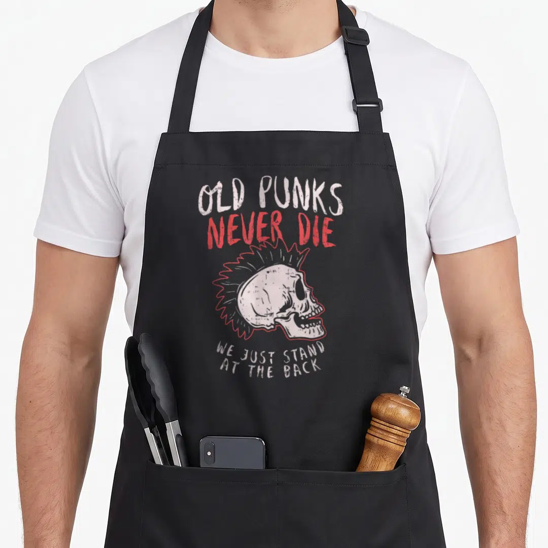 Die punk rock apron - Mohawk skull with Old Punks Never Die text in red and grey distressed font — shown on a black cotton canvas apron against a white background