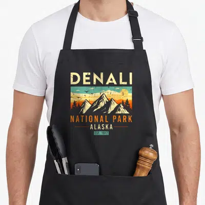 Park alaska apron - Retro Denali National Park graphic with mountains, sun, birds, and cream orange teal yellow text — shown on a black cotton canvas apron against a white background