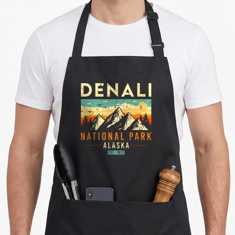 Park alaska apron - Retro Denali National Park graphic with mountains, sun, birds, and cream orange teal yellow text — shown on a black cotton canvas apron against a white background