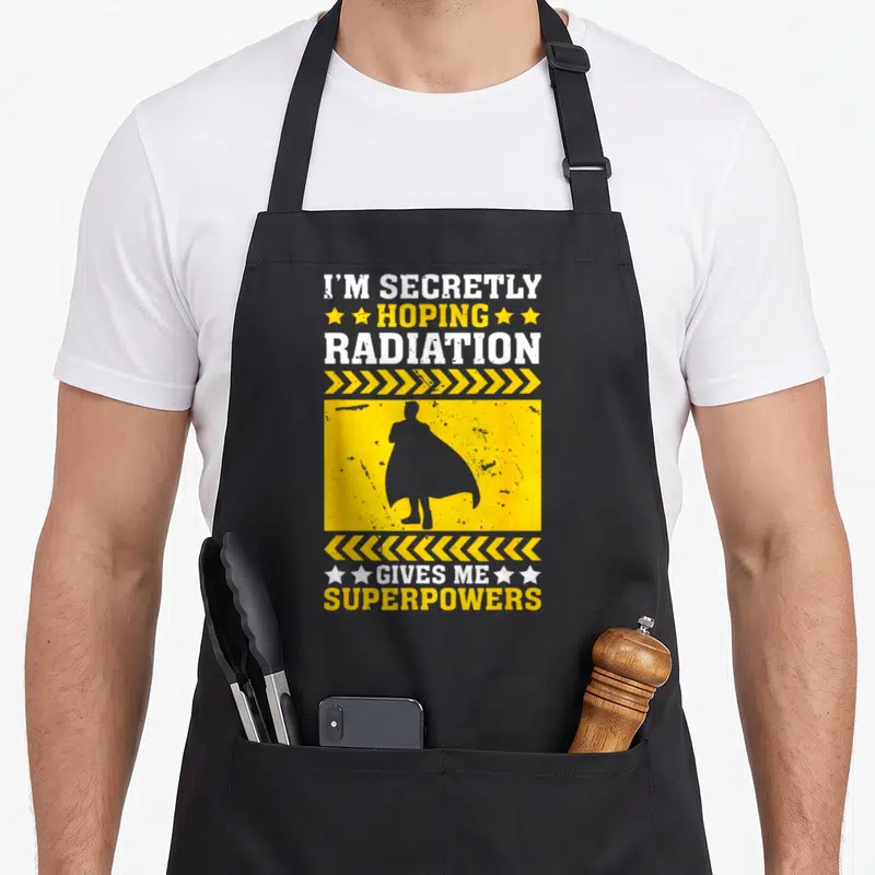 Awareness radiation aprons - I'M SECRETLY HOPING RADIATION GIVES ME SUPERPOWERS text with superhero silhouette — shown on a black cotton canvas apron against a white background
