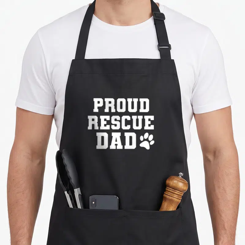 Proud grill rescue apron - White PROUD RESCUE DAD text with paw print icon — shown on a black cotton canvas apron against a white background