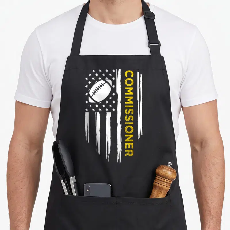 Commissioner american flag apron - Distressed white flag with football and vertical yellow COMMISSIONER text — shown on a black cotton canvas apron against a white background