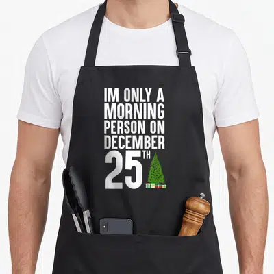 December 25th women's christmas apron - White stacked text next to green Christmas tree and gifts on black apron — shown on a black cotton canvas apron against a white background