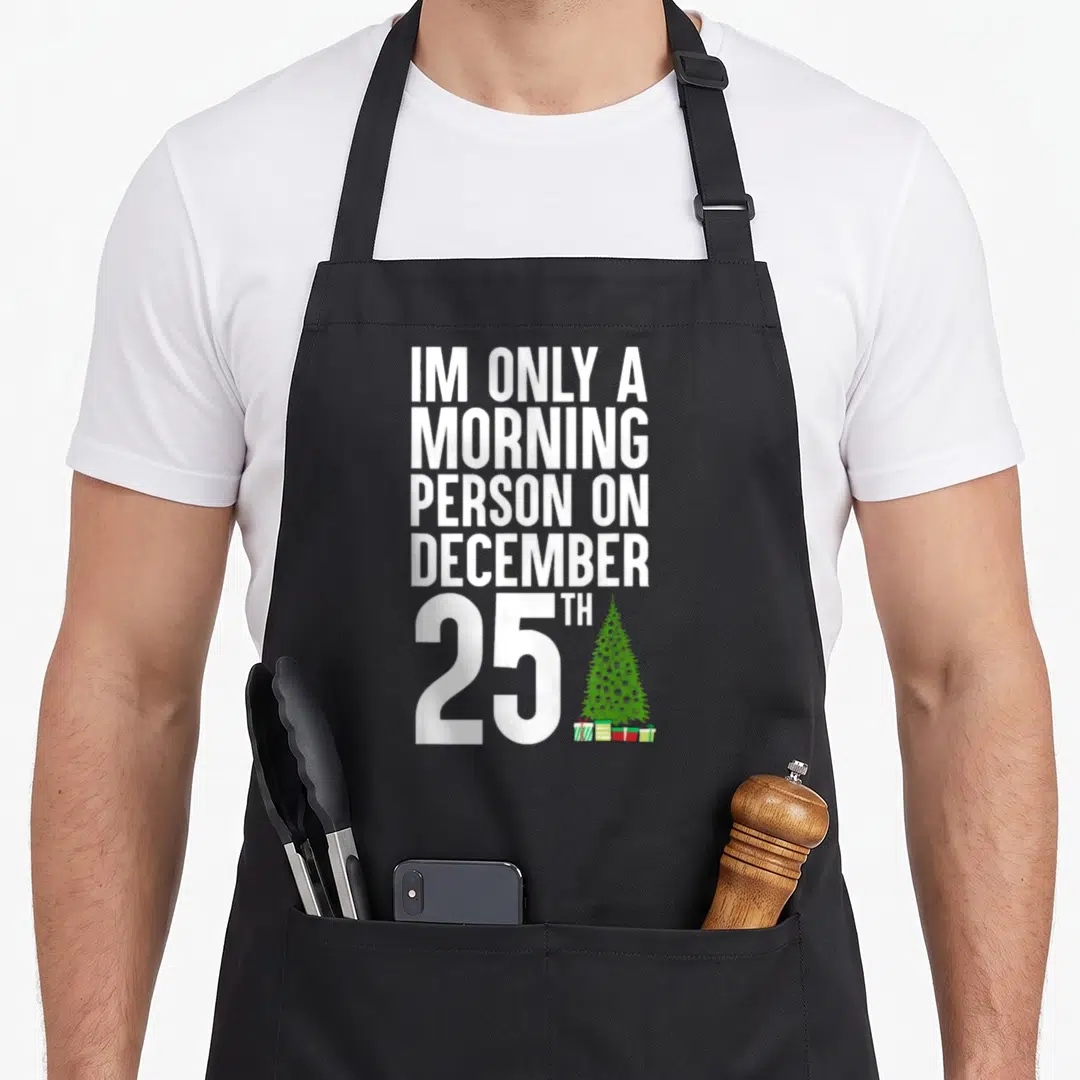 December 25th women's christmas apron - White stacked text next to green Christmas tree and gifts on black apron — shown on a black cotton canvas apron against a white background