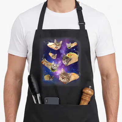 Taco space cat apron - Six cats wrapped in tacos floating in a purple and blue starry space background — shown on a black cotton canvas apron against a white background