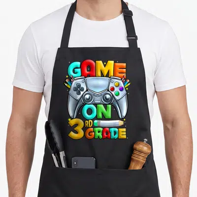 Grade game aprons - Central controller with GAME ON 3RD GRADE text and scattered pencils paintbrushes stars in cyan red — shown on a black cotton canvas apron against a white background
