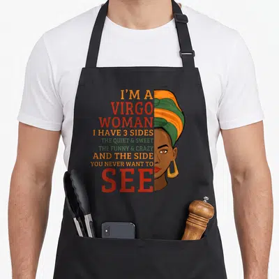 Cute virgo apron - I'M A VIRGO WOMAN text and cartoon woman face with headwrap in red orange green and brown — shown on a black cotton canvas apron against a white background