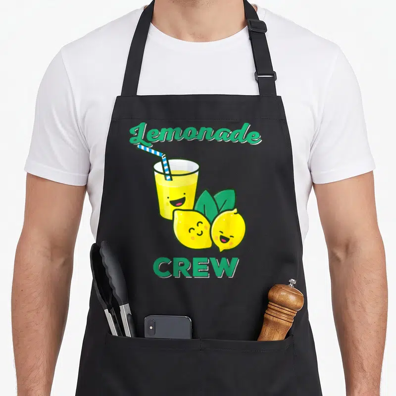 Lemonade lemon apron - Cartoon lemonade glass with straw and two smiling lemons with green text — shown on a black cotton canvas apron against a white background