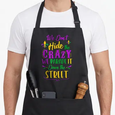 Hide mardi gras aprons - Stacked text We Don't Hide the Crazy WE PARADE IT Down the Street in purple yellow green — shown on a black cotton canvas apron against a white background
