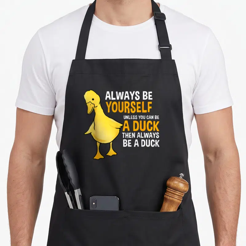 Alway duck apron - Yellow cartoon duck standing next to stacked white and orange text reading Always Be Yourself Unless — shown on a black cotton canvas apron against a white background