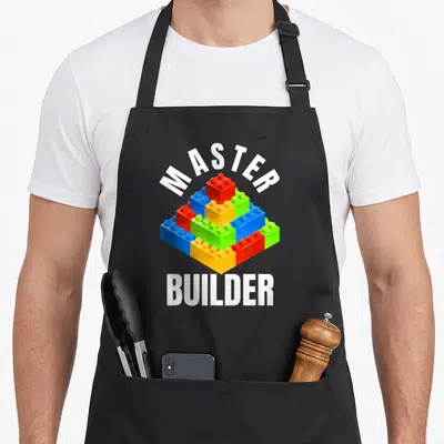 Master builder block & apron - Pyramid of white red blue yellow and green building blocks with master builder text — shown on a black cotton canvas apron against a white background