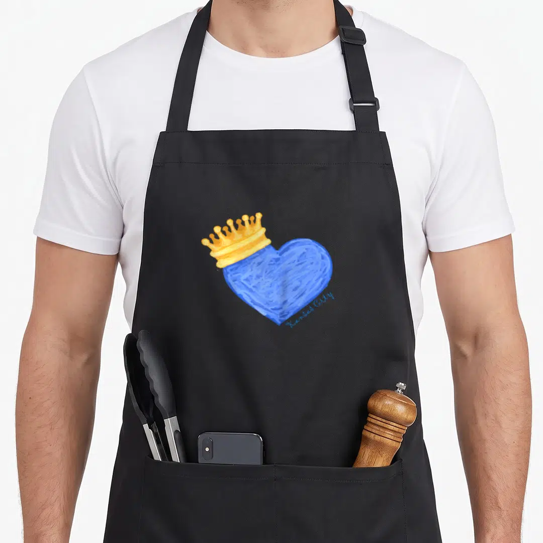Kansas crown royal apron - Blue heart illustration topped with a gold crown and small script reading Kansas City — shown on a black cotton canvas apron against a white background