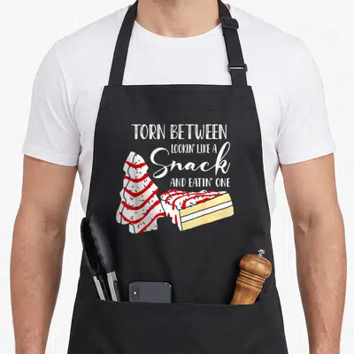 Christmas cake apron - Tree-shaped cake slice in white red yellow with text torn between snack and eatin one — shown on a black cotton canvas apron against a white background