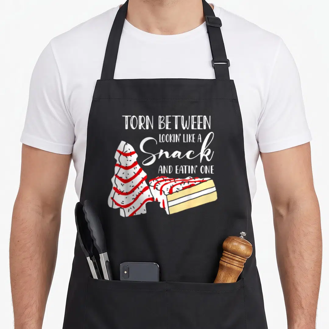 Christmas cake apron - Tree-shaped cake slice in white red yellow with text torn between snack and eatin one — shown on a black cotton canvas apron against a white background