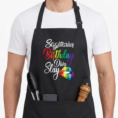 November rainbow apron - Sagittarius Birthday Diva Slay text with rainbow colored lips on black background — shown on a black cotton canvas apron against a white background