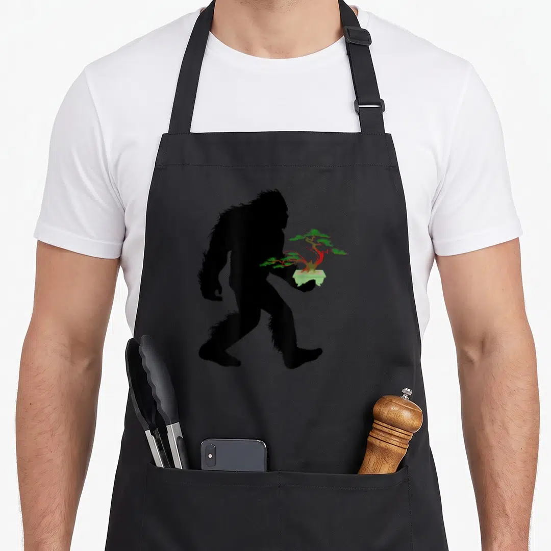 Carrying tree apron - Black Bigfoot silhouette walking right holding a green and red bonsai tree in a pot — shown on a black cotton canvas apron against a white background
