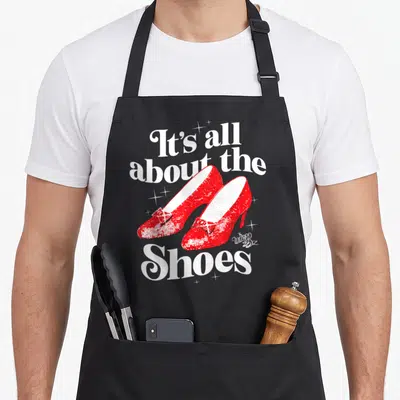 Gale dorothy apron - Two red ruby slippers with white text It's all about the Shoes and scattered sparkles — shown on a black cotton canvas apron against a white background