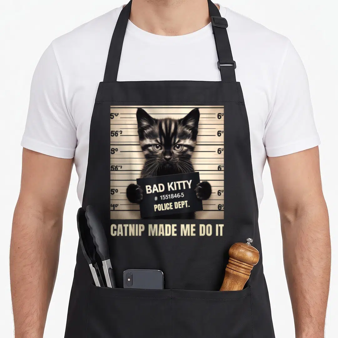 Catnip cat dad apron - Kitten mugshot holding bad kitty sign with catnip made me do it caption — shown on a black cotton canvas apron against a white background