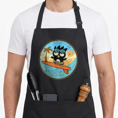 Badtz surfing apron - Cartoon black penguin on orange surfboard inside circular beach scene with palm tree and sun — shown on a black cotton canvas apron against a white background