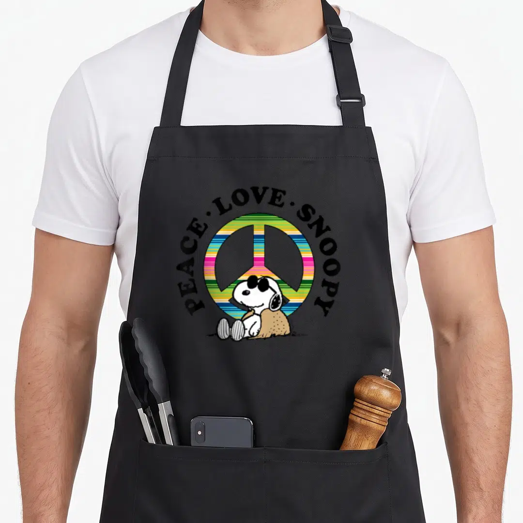 Funny peace apron - Snoopy sitting before multicolored peace symbol with arched text PEACE LOVE SNOOPY — shown on a black cotton canvas apron against a white background