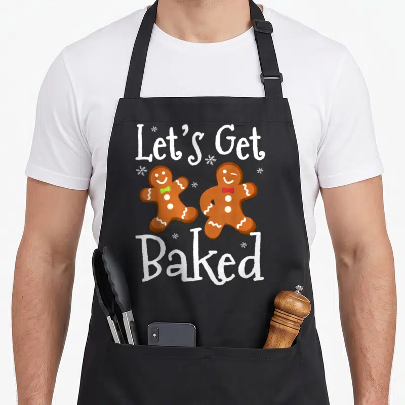 Baked christmas cookie baking team aprons - Two gingerbread men with bowties under Let's Get Baked text and snowflakes — shown on a black cotton canvas apron against a white background