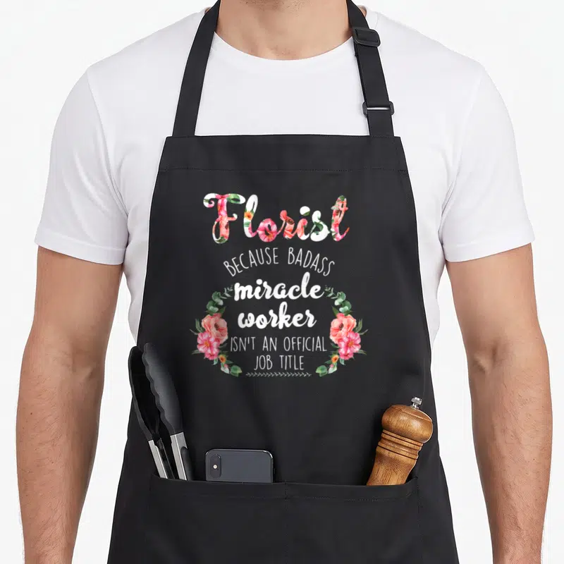Hard gardener apron - Florist text filled with pink watercolor flowers and white stacked quote flanked by two wreaths — shown on a black cotton canvas apron against a white background