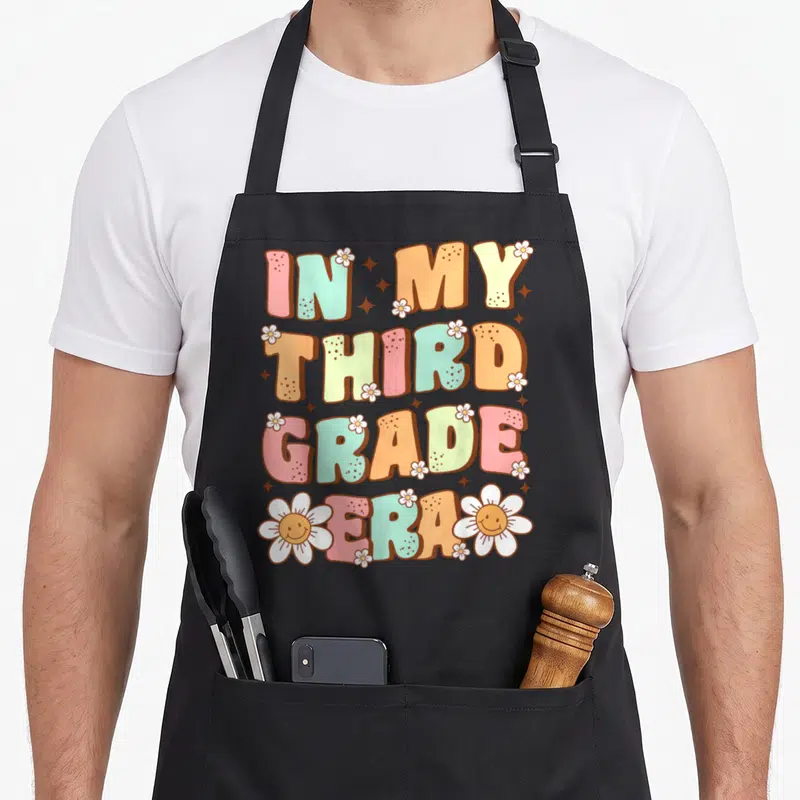 Third grade era apron - Stacked retro text IN MY THIRD GRADE ERA with pink orange green yellow white letters — shown on a black cotton canvas apron against a white background