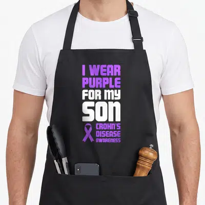 Son crohns awareness apron - Purple and white stacked text I WEAR PURPLE FOR MY SON CROHN'S DISEASE AWARENESS — shown on a black cotton canvas apron against a white background