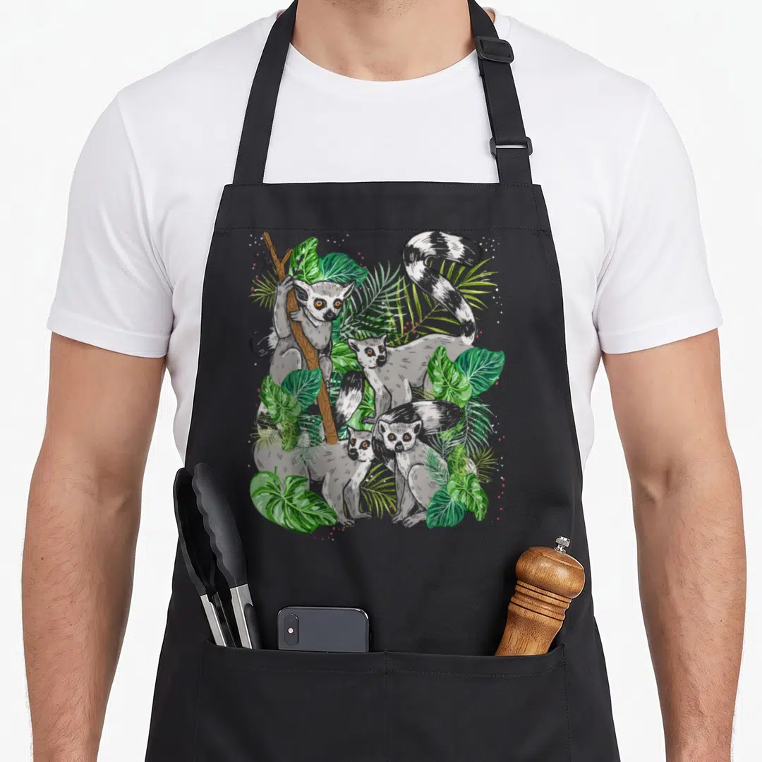 Tropical animal apron - Three grey lemurs clustered among green tropical leaves and a brown branch — shown on a black cotton canvas apron against a white background