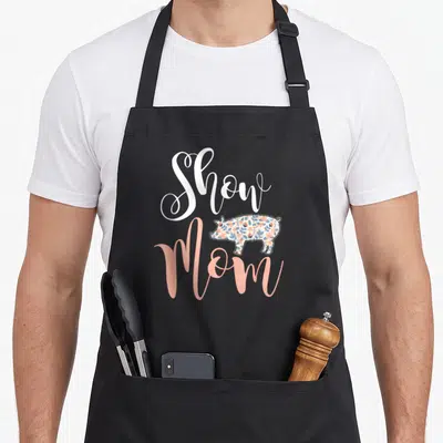 Show cooking mama apron - White Show Mom script with peach and blue floral pig illustration — shown on a black cotton canvas apron against a white background