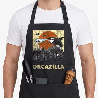 Orcazilla orca dad apron - Giant orca firing beam at helicopters above city skyline with ORCAZILLA text — shown on a black cotton canvas apron against a white background