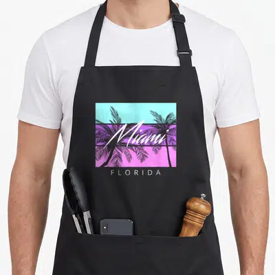 Souvenir 80s apron - White Miami script over purple palms and cyan purple pink stripes with Florida text below — shown on a black cotton canvas apron against a white background