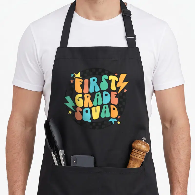 School groovy teacher apron - Bubbly multicolored text FIRST GRADE SQUAD over black white checkerboard circle — shown on a black cotton canvas apron against a white background