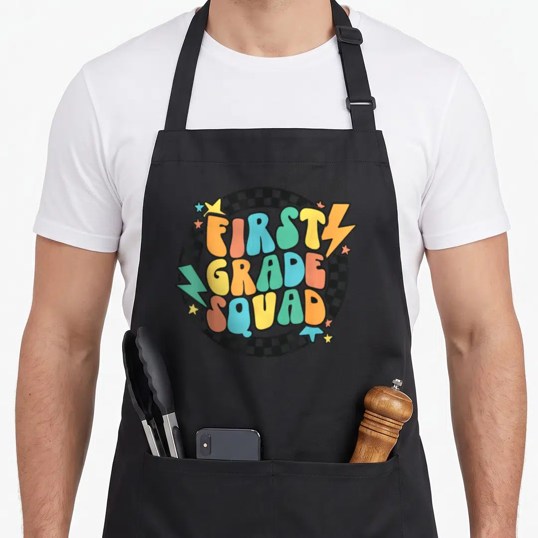 School groovy teacher apron - Bubbly multicolored text FIRST GRADE SQUAD over black white checkerboard circle — shown on a black cotton canvas apron against a white background
