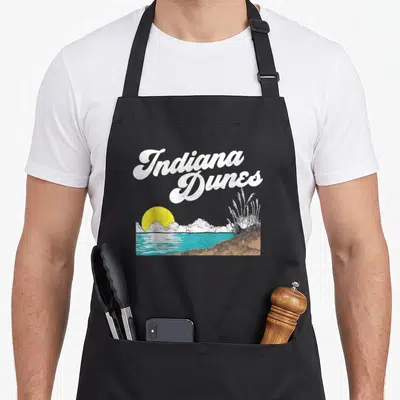 Indiana 80s apron - Retro Indiana Dunes landscape print with white script, yellow sun, turquoise water, and tan dunes — shown on a black cotton canvas apron against a white background