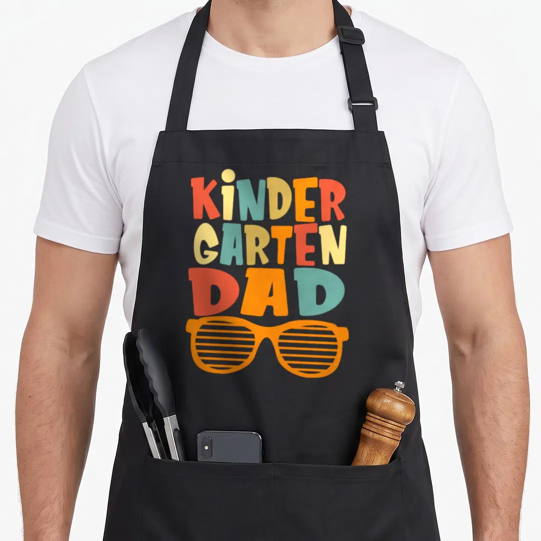 Kindergarten school dad apron - Stacked text reading kindergarten dad in red orange blue and yellow above a pair — shown on a black cotton canvas apron against a white background