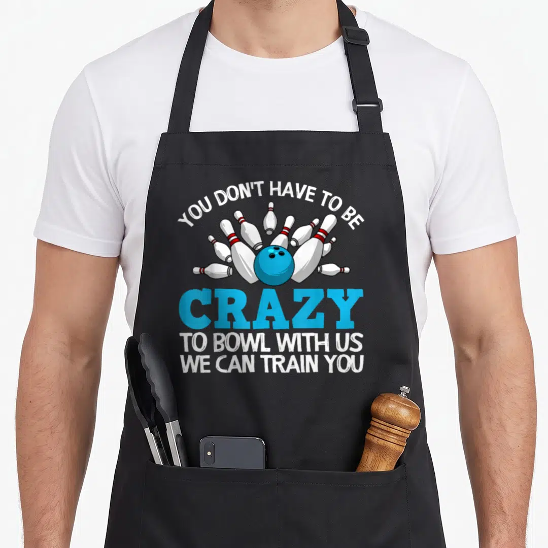 Bowling pins for aprons - Blue bowling ball scattering ten white pins with red stripes and bold text — shown on a black cotton canvas apron against a white background