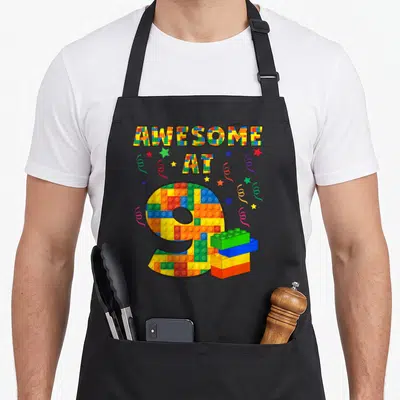 Age building aprons - Awesome At 9 printed in colorful building blocks with stars and streamers — shown on a black cotton canvas apron against a white background