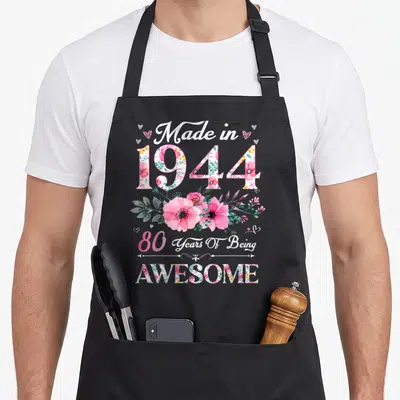 Cute 80th vintage floral apron - Watercolor floral 1944 and 80 Years Of Being Awesome text design with hearts — shown on a black cotton canvas apron against a white background