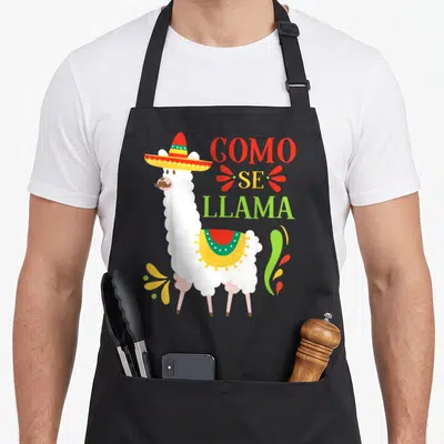 Como mexican apron - White llama in sombrero with stacked COMO SE LLAMA text and colorful swirls — shown on a black cotton canvas apron against a white background