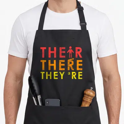 Lesson teacher apron - Stacked text THEIR, THERE, THEY'RE with person icon replacing I and arrow replacing E in red — shown on a black cotton canvas apron against a white background