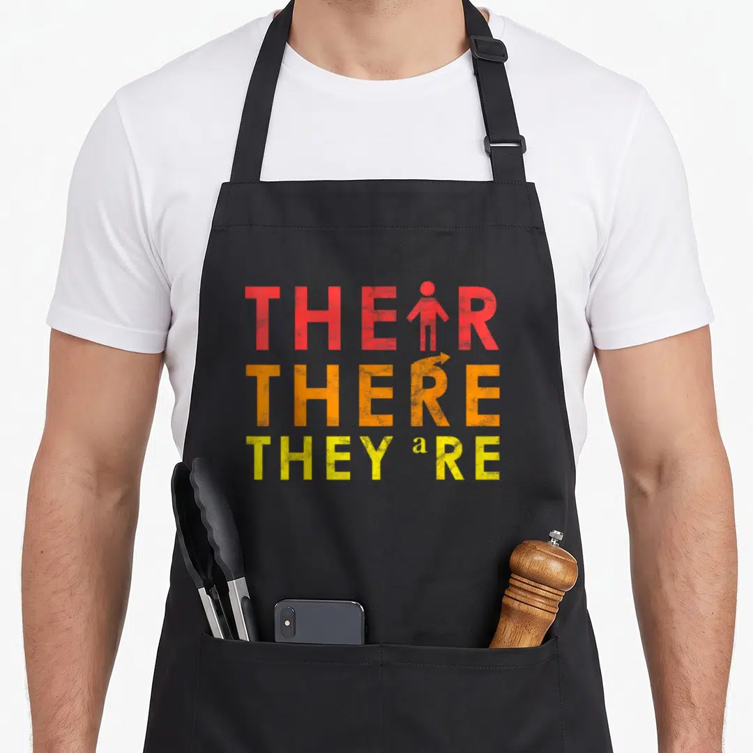 Lesson teacher apron - Stacked text THEIR, THERE, THEY'RE with person icon replacing I and arrow replacing E in red — shown on a black cotton canvas apron against a white background